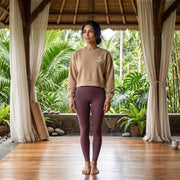 A woman stands barefoot in a wooden pavilion wearing a beige cropped sweatshirt with a small white chest graphic and mauve-purple leggings, with lush tropical plants visible in the background.