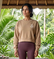 A woman stands under a thatched roof in a tropical garden setting, wearing a beige cropped sweatshirt with a small white graphic on the left chest and mauve-purple leggings.