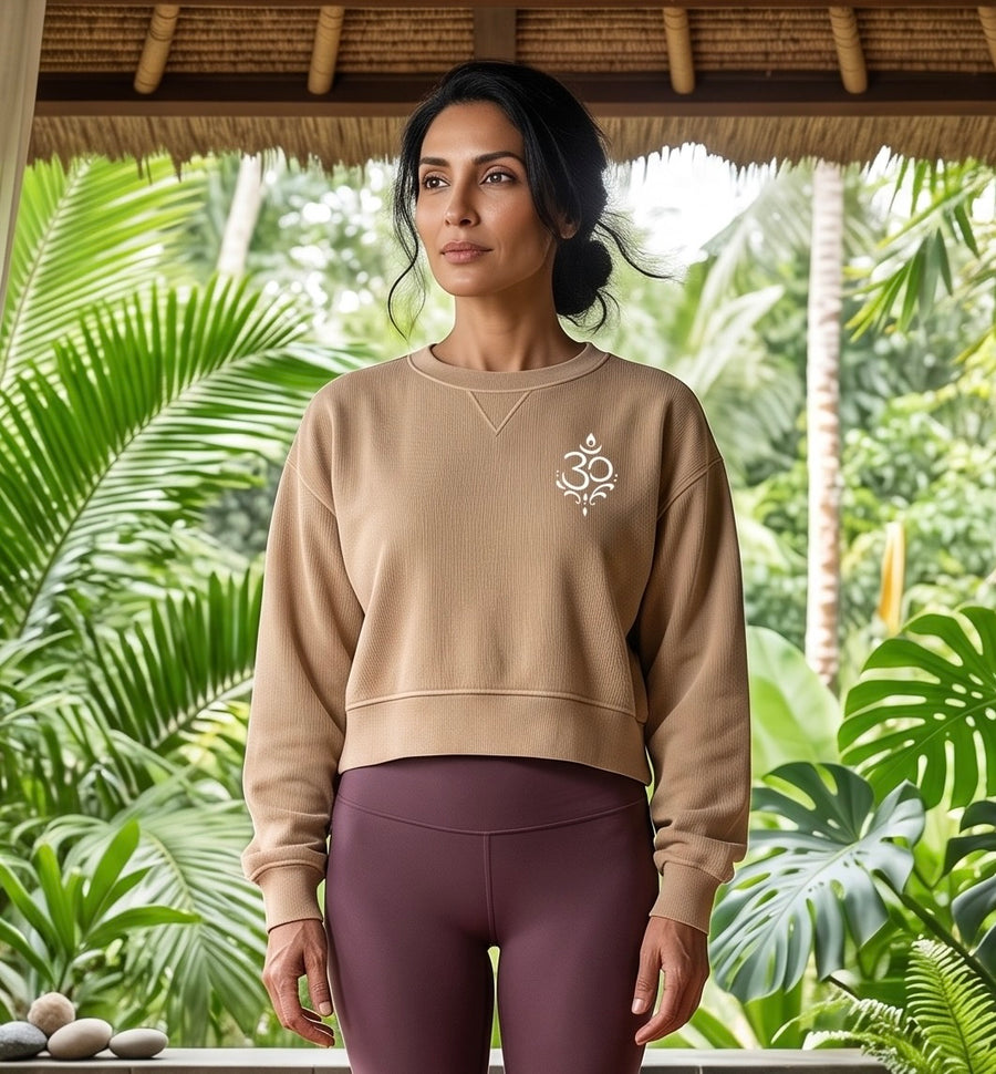 A woman stands under a thatched roof in a tropical garden setting, wearing a beige cropped sweatshirt with a small white graphic on the left chest and mauve-purple leggings.