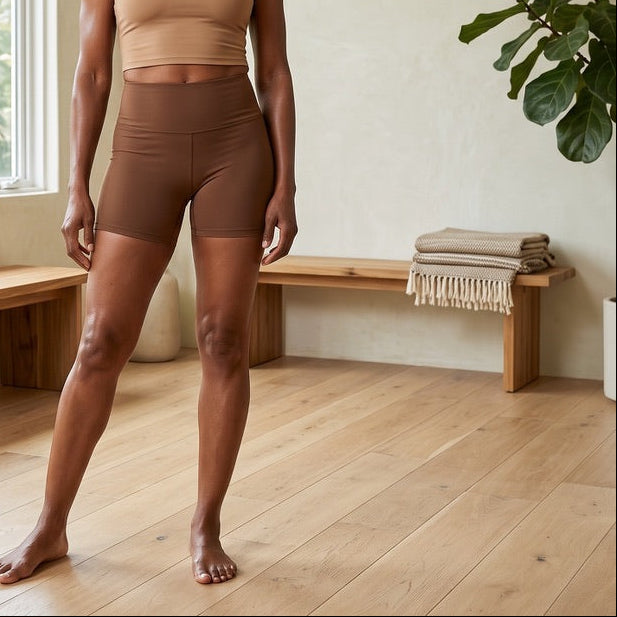 A woman stands barefoot in a bright, minimalist room wearing a beige sports bra and brown fitted biker shorts. Wooden benches, a rolled yoga mat, and a potted plant are visible in the background.