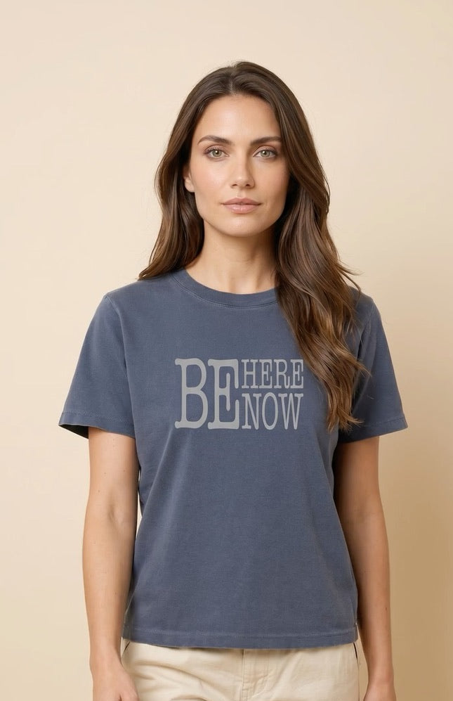 Woman wearing a blue t-shirt with 'BE HERE NOW' text on a beige background