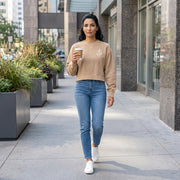 A woman walking on a city sidewalk wearing a beige crewneck sweatshirt with a small white graphic on the left chest, blue jeans, and white sneakers, while holding a paper coffee cup.