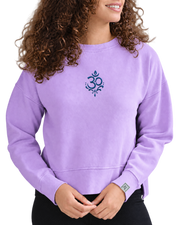 A woman wearing a lavender crewneck sweatshirt with a blue Om symbol printed on the center chest, paired with black pants, against a plain white background.