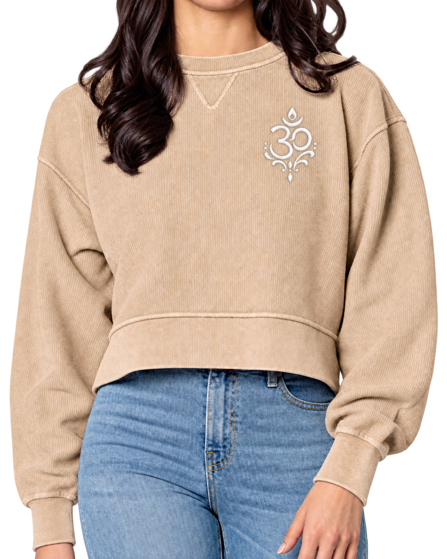 A woman wearing a cropped beige sweatshirt with a white Om symbol on the left chest, paired with blue jeans, shown against a plain white background.